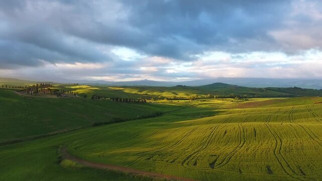 Aerial view of rolling green hills under a dramatic cloudy sky at sunset in Tuscany, Italy.