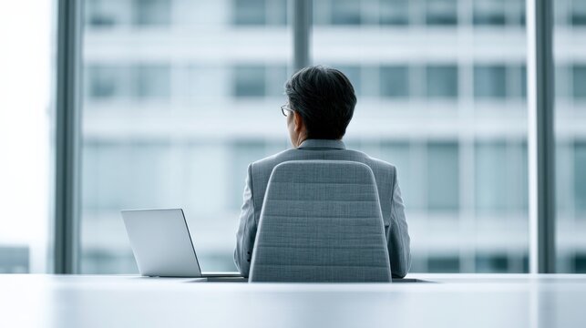 A businessman sits at a desk in a modern office, looking out of the window at a cityscape. A laptop is open on the desk before him.
