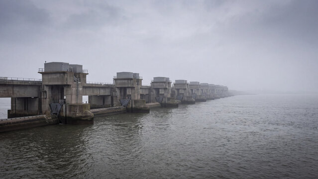 Concrete tidal barrage with sluice gates stretching across estuary in thick fog. Storm surge barrier and flood defense dam for coastal protection, water management and tidal power generation