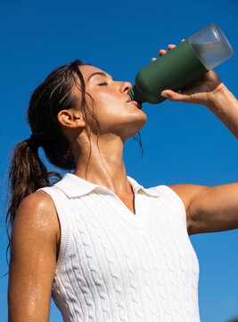 Athletic young woman drinking water from bottle after workout against clear blue sky.