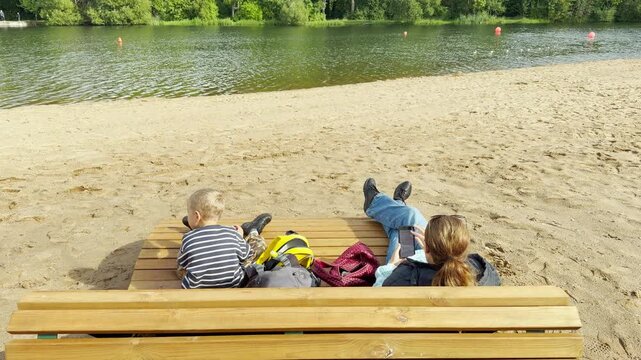 A mother and child relax on a wooden bench at Kolonistsky Pond Beach in Pushkin, Saint Petersburg. This peaceful scene feels calm and cozy. They enjoy a quiet moment by the water.