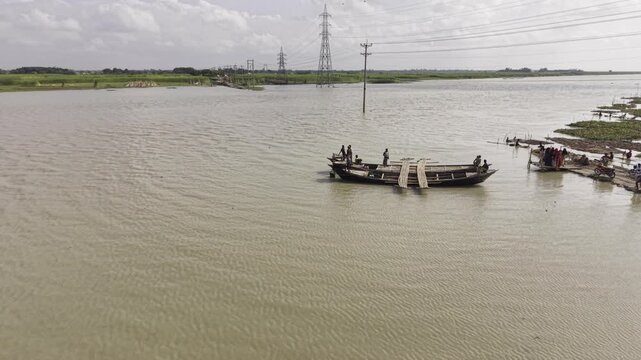 Drone footage shows wooden boats on the wide Padma River, with calm muddy water, green riverbanks, distant power transmission towers and open rural landscape under a bright cloudy sky.