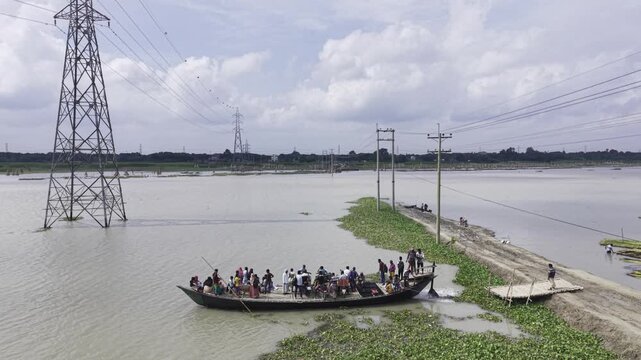 Aerial drone footage shows a crowded passenger boat crossing the Padma River in Bangladesh, with a narrow riverside embankment, transmission towers, floating vegetation, blue sky and white clouds.