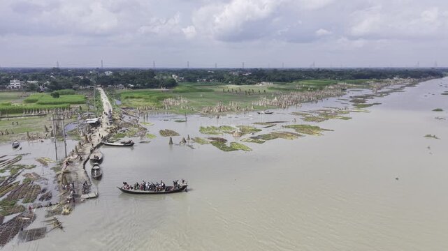 Drone aerial footage of a busy passenger boat on the Padma River in Bangladesh, showing riverside vegetation, an embankment, power transmission towers, blue sky and white clouds.