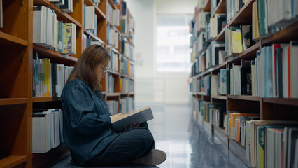 A woman sits cross-legged on the floor between tall library bookshelves filled with books, quietly reading an open book in her hands at library © M Stocker