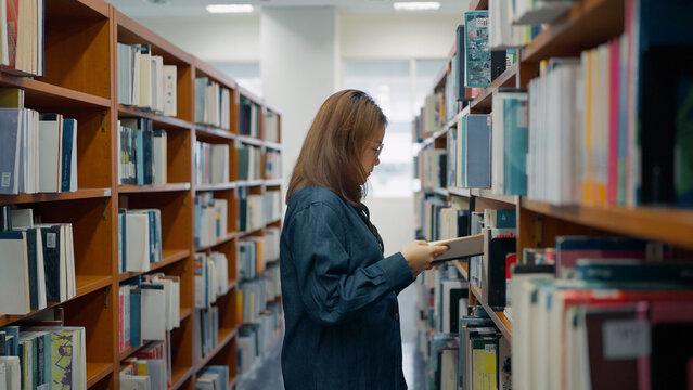 A woman reading a book and standing in between several bookshelves that are full of books at an library