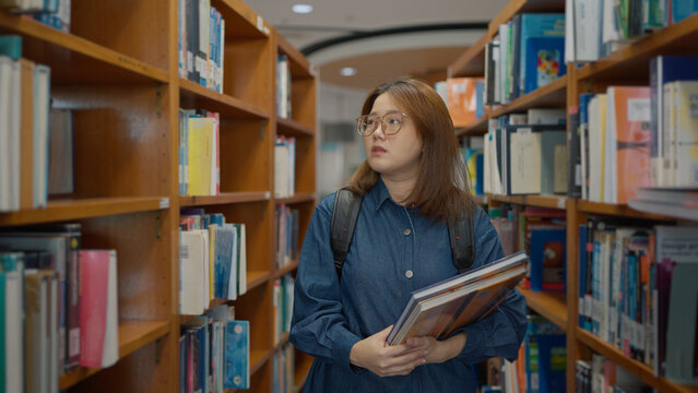 A young woman with glasses and a backpack stands in a library aisle holding books, looking thoughtfully at the shelves around her at library