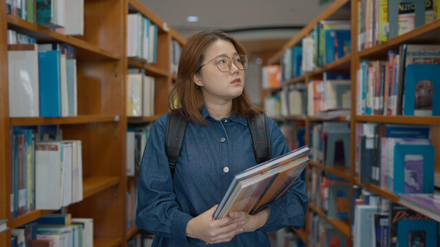 A young woman with glasses and a backpack stands in a library aisle holding books, looking thoughtfully at the shelves around her at library