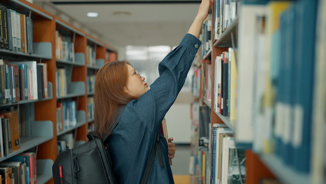 A young woman with a backpack reaches up to pull a book from a high shelf in a quiet library aisle filled with books