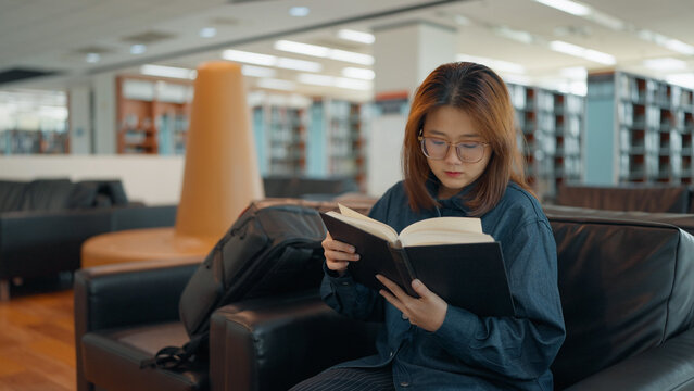 A young woman wearing glasses sits on a comfortable sofa in a spacious library, focused on reading a book held in her hands