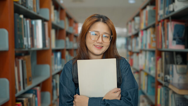 A smiling woman with glasses holds a book to her chest while standing between tall, book-filled library shelves