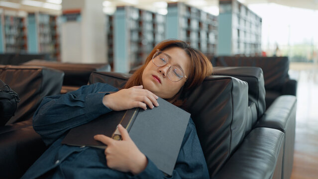 A young woman wearing glasses sleeps peacefully on a sofa in a library, hugging a dark book tightly against her chest while taking a break