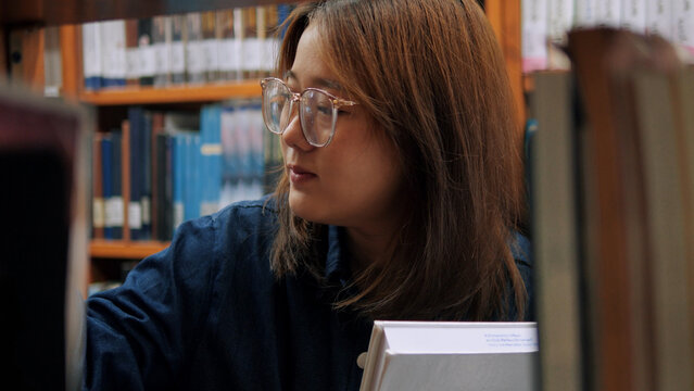 A young woman with glasses looking carefully at books on library shelves while holding a book in her hands