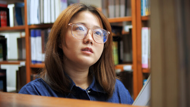 A portrait of a young woman with glasses looking up thoughtfully between wooden library bookshelves filled with books