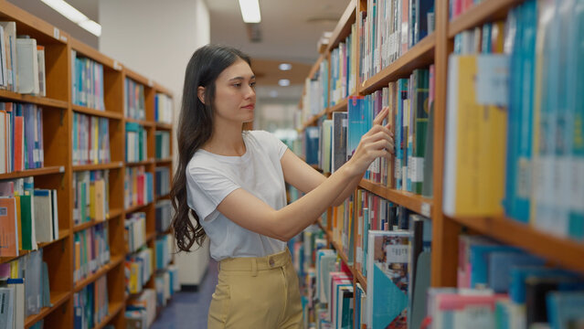 A young woman in a white t-shirt stands in a library aisle, reaching out to select a book from a wooden shelf filled with books