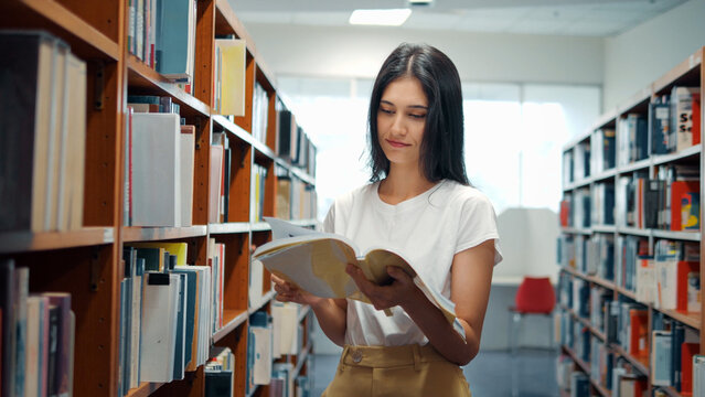 A young woman in a white t-shirt stands in a library aisle, reading an open book held in both hands between rows of wooden shelves