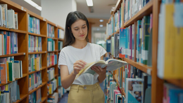 A young woman in a white t-shirt stands between library bookshelves, holding an open book while reading and flipping through its pages carefully