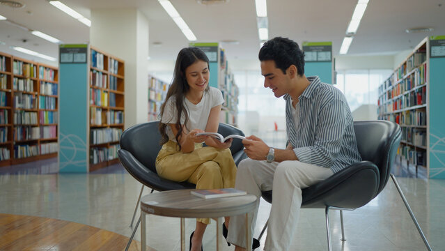 A young woman and a man sit together in modern library armchairs, smiling as they look at an open book, with bookshelves surrounding them