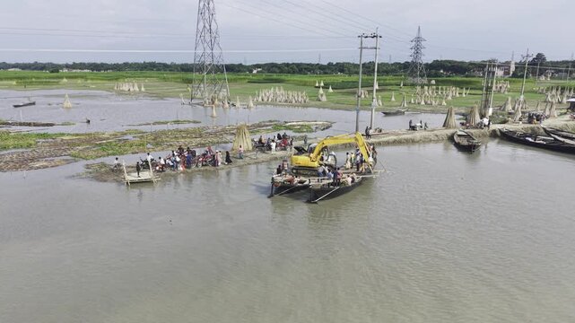 Bangladesh, 15 August 2026: Aerial drone footage shows an excavator barge crossing the Padma River near a rural village, with passenger boats, people, floating vegetation, power lines and green fields