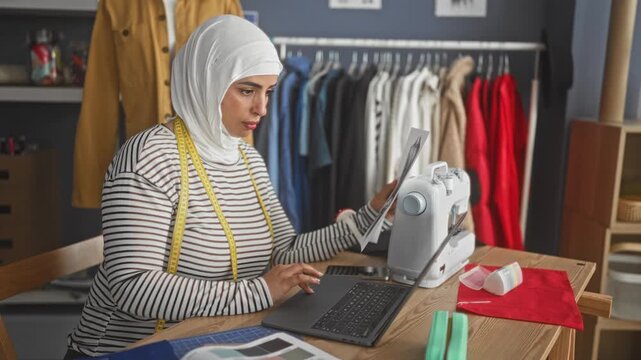 Woman in hijab holding a sewing pattern and typing on laptop beside sewing machine in studio; concentration craft entrepreneurship.