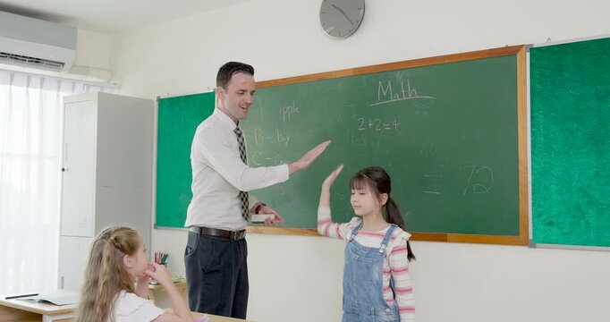 Male primary teacher gives high five to young girl near chalkboard, then writes math notes as pupils watch, creating back to school confidence, class rule practice, and positive lesson guidance.
