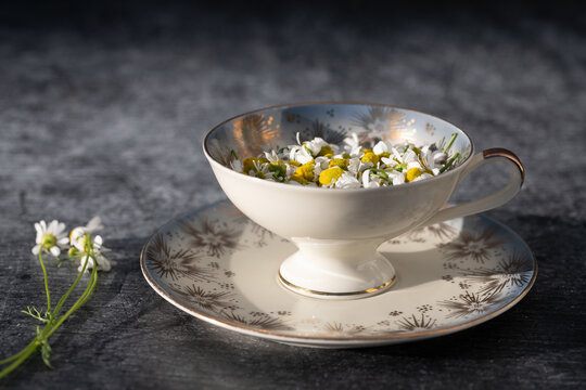 A close-up of an old teacup filled with chamomile flowers. A dark table is visible in the background.