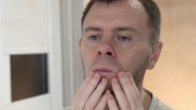 Close-up of adult man using hands to apply skincare on cheeks and jaw while looking into bathroom mirror with focused grooming expression and facial massage gestures