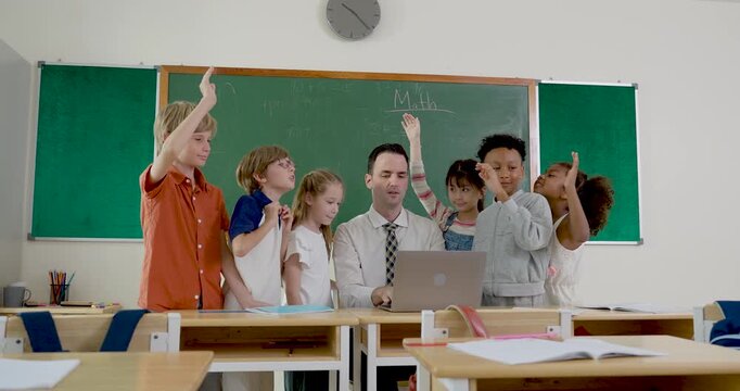 Diverse elementary pupils celebrate around male teacher at laptop, raising arms and smiling after successful class task, building peer teamwork, confidence, motivation, and school friendship.