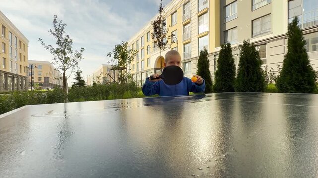 A focused young boy swings a paddle while playing table tennis. He stands behind a table in a bright outdoor courtyard near modern buildings. His eager look shows pure excitement.