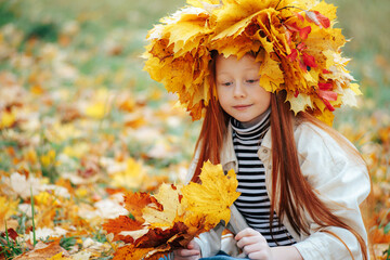 Close up of a redhead girl in a leaf crown sitting on the ground looking down at a yellow maple bouquet. © Dmytro