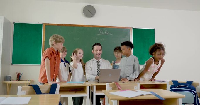 Male adult teacher guides young multicultural girl and boy student around laptop during back to school elementary class activity, building teamwork, question time, notebook practice and lesson routine
