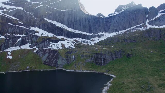 Stunning aerial footage flying towards a majestic mountain range with lingering snow patches and a serene lake in the Lofoten Islands, Norway