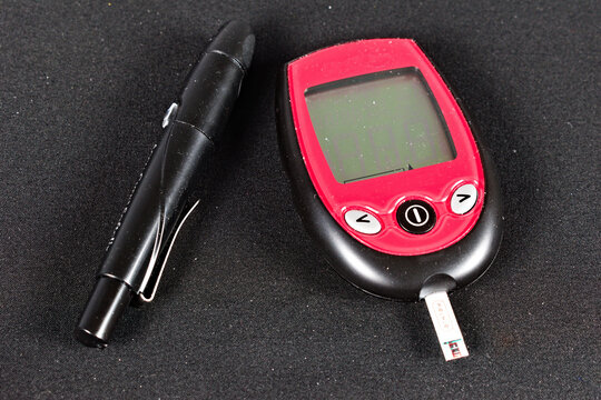 A red and black glucose meter with a test strip inserted sits next to a black lancet pen on a textured dark surface.