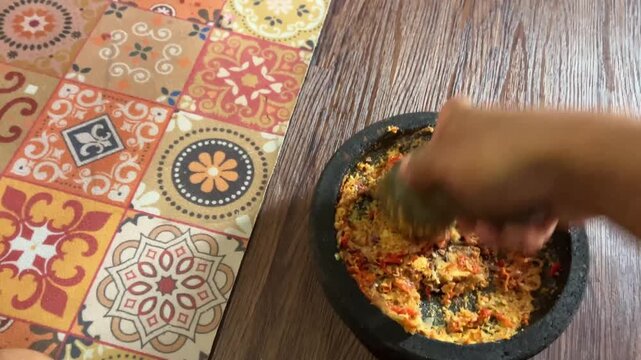 Slow motion first person POV video of hands grinding and mashing spices with a stone mortar and pestle on a patterned mat for traditional cooking preparation.