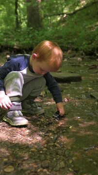 Vertical screen. Young child with red hair crouches near a stream, extending a hand into the water to touch stones and pebbles, with vibrant green trees and sunlight creating a serene outdoor setting