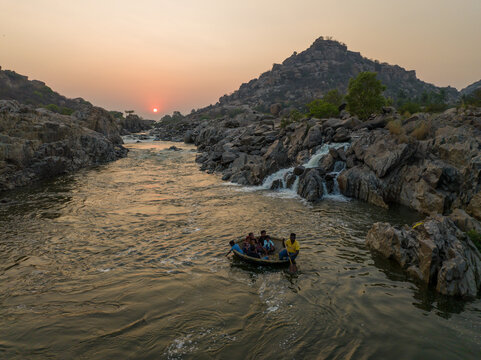 Aerial view of a coracle boat carrying people on the Tungabhadra River surrounded by rocky terrain and boulders during sunset in Hampi, Karnataka, India.