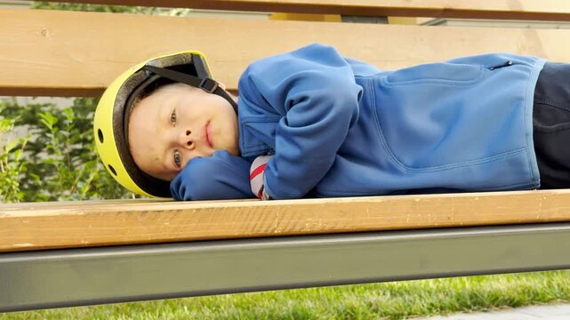 A tired young boy lies down to rest on a wooden bench outdoors. Wearing a yellow helmet, he looks exhausted yet calm in the park. The sleepy child feels cozy and relaxed.