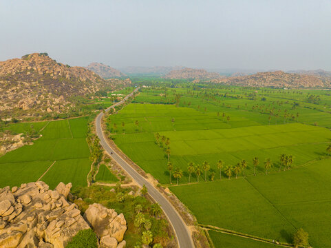 Aerial view of the lush green rice paddies and winding road surrounded by ancient rocky hills in the historic landscape of Hampi, Karnataka, India.
