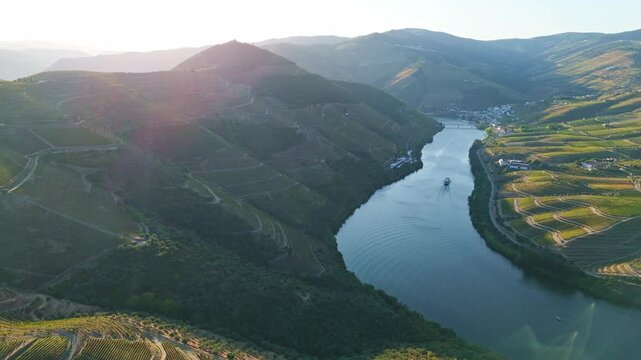 Drone view of Douro Valley, Portugal, with the Douro River, vineyards, tourist boat, and sunset reflection with bright river reflection and distant hills. Pan shot