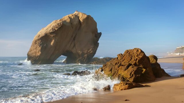 Penedo do Guincho stands tall at Praia de Santa Cruz in Portugal. Ocean waves crash against the sandy beach as the sun sets, casting shadows on the rock arch and creating a beautiful view