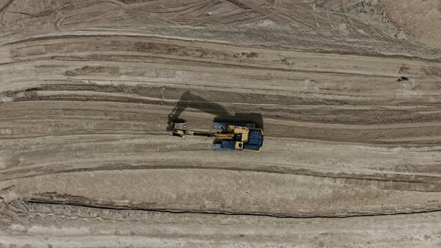 Aerial view of a yellow excavator working on a large dirt construction site with visible tire tracks in the soil in Longmont, Colorado, United States.