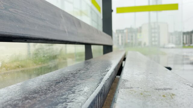 A wooden bench sits wet with raindrops inside an empty bus stop shelter. The blurry street scene outside creates a gloomy, lonely, and melancholic mood.