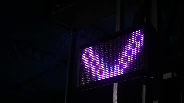 A bright purple LED digital display board showing a downward arrow symbol in the dark.