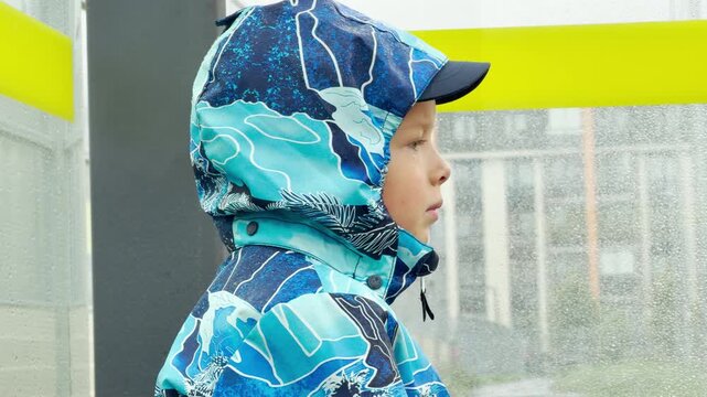 A young boy in a blue patterned raincoat stands thoughtfully inside a wet bus shelter. The rainy weather creates a quiet, lonely, and melancholic mood.