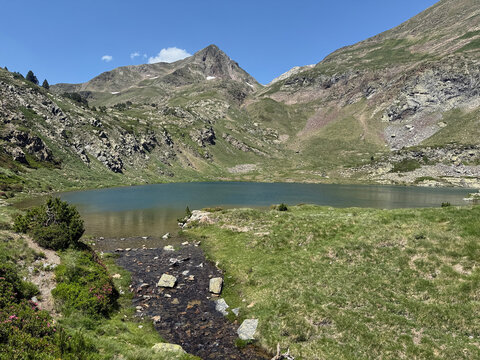 A scenic view of a high-altitude mountain lake fed by a small stream in the Pyrenees. The landscape features rocky peaks and green alpine meadows under a clear blue sky.