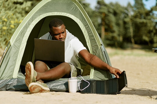 Young adult African American Black man sitting in camping tent using laptop connected to portable solar panel charger. Concept for remote work, sustainable energy, digital nomad marketing