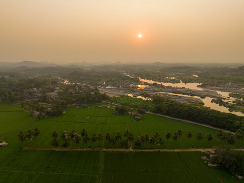 Aerial view of the historic Hampi landscape featuring lush green rice paddies, rows of palm trees, and the winding Tungabhadra river under a golden sunset in Hampi, Karnataka, India.
