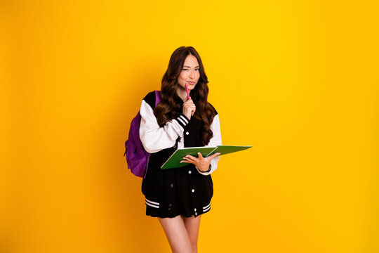 Thoughtful female student with purple backpack and green notebook studying and smiling in a varsity jacket against a bright yellow background