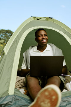 Vertical shot of happy young African American man sitting in camping tent using laptop computer. Concept represents digital nomad lifestyle for remote work campaigns