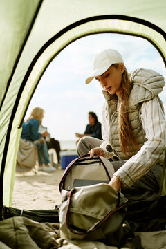Vertical shot of young Caucasian woman unpacking backpack outside camping tent with friends resting in background. Image suited for outdoor adventure gear advertising